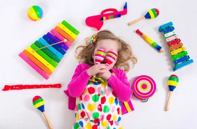 Child lying on floor holding maracas, surrounded by colorful musical toys and instruments