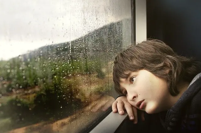 Child rests head by rain-covered window, looking outside with thoughtful, quiet expression