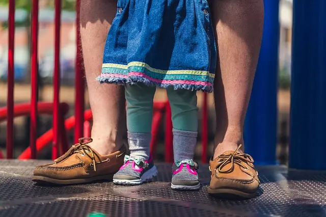 Child stands between an adult’s legs on playground platform, wearing dress and trainers