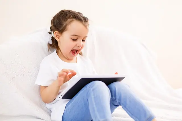 Child sitting on sofa, smiling at a tablet with excited expression