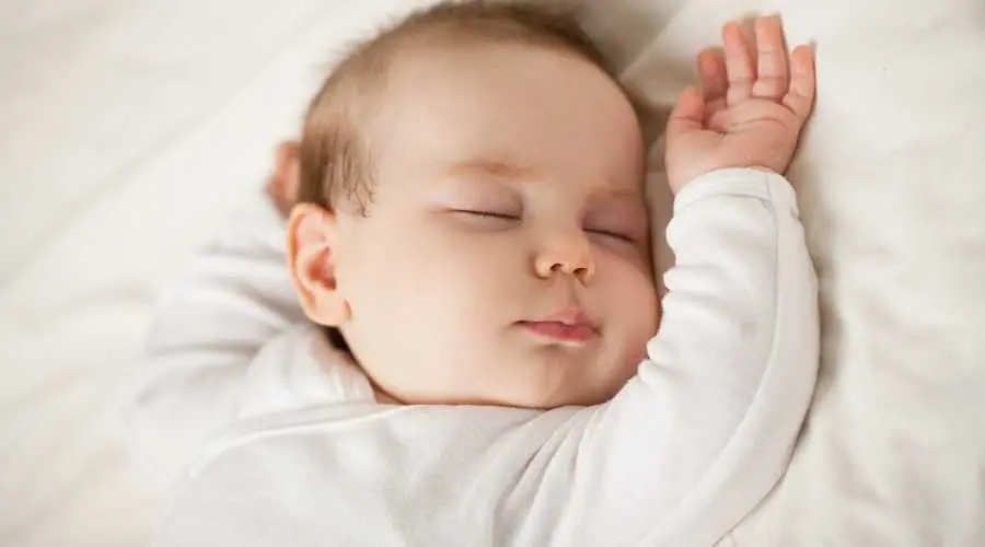 Sleeping baby in white outfit with one arm raised on a soft white bed