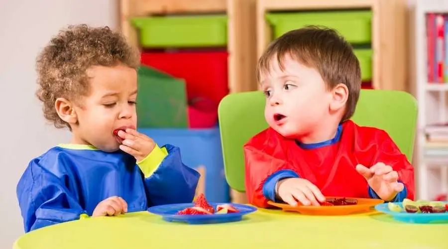 Two children eating fruit at a table, wearing bibs in a classroom