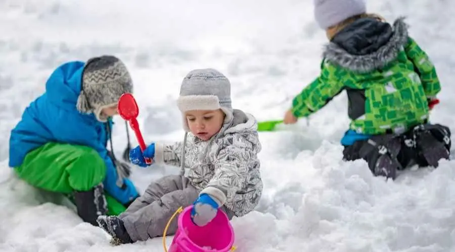 Three children in winter clothes play in the snow with buckets and shovels