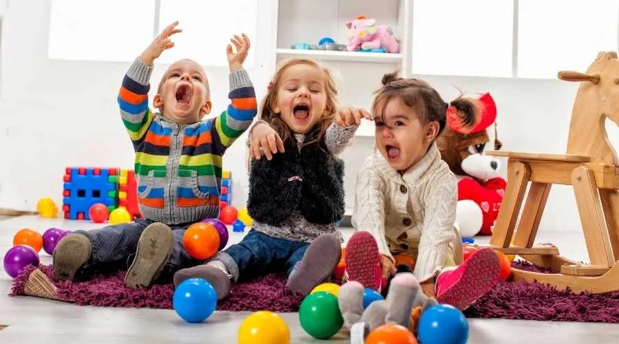 Three children sit on a rug in a playroom, laughing among colourful balls.
