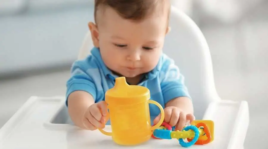 Baby in high chair reaching for a yellow cup and colourful teething toy