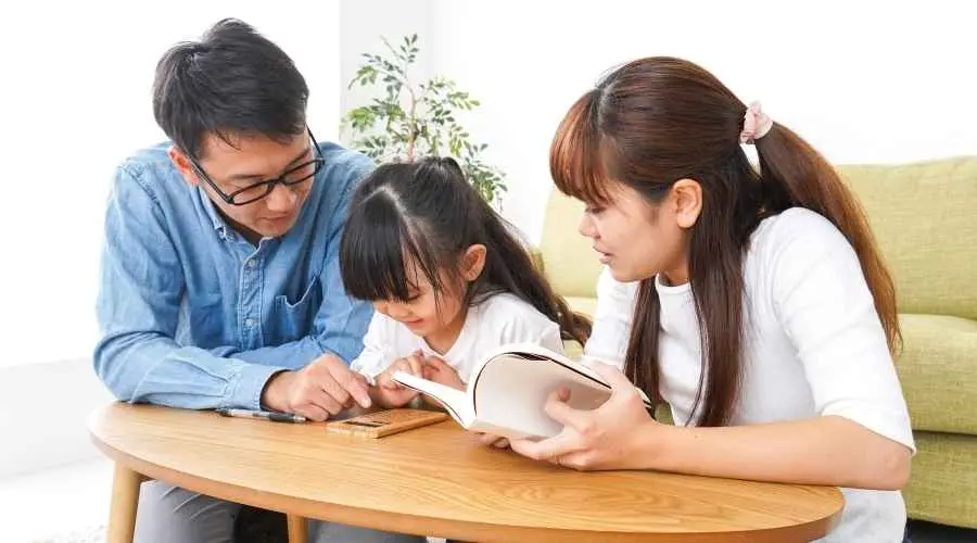 Two adults and a child read together at a small table in a bright living room.