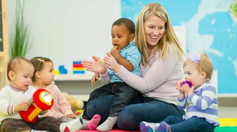 Adult sits with three toddlers on a mat, clapping in a bright classroom.