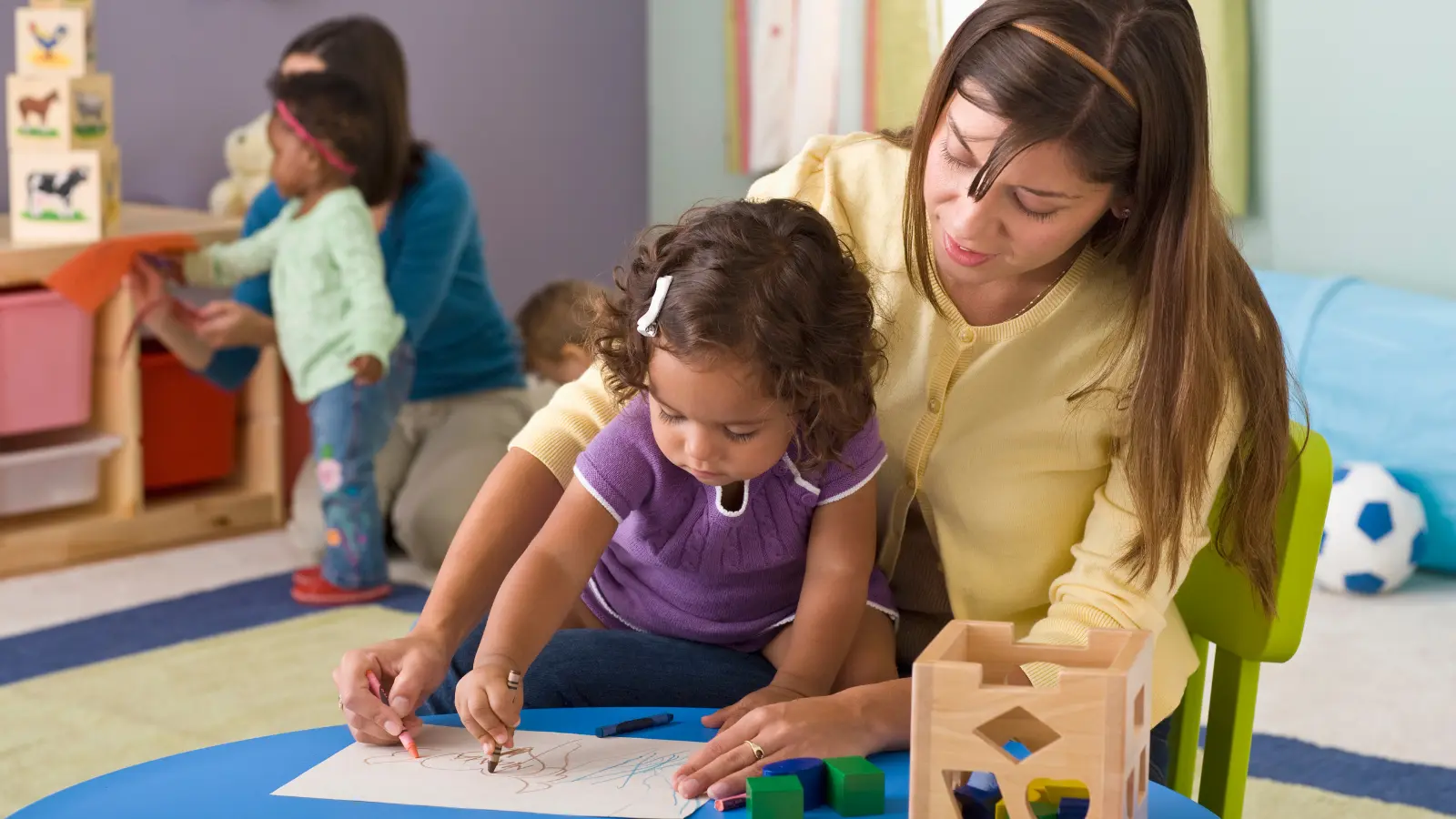 Young girl drawing with crayons alongside a teacher