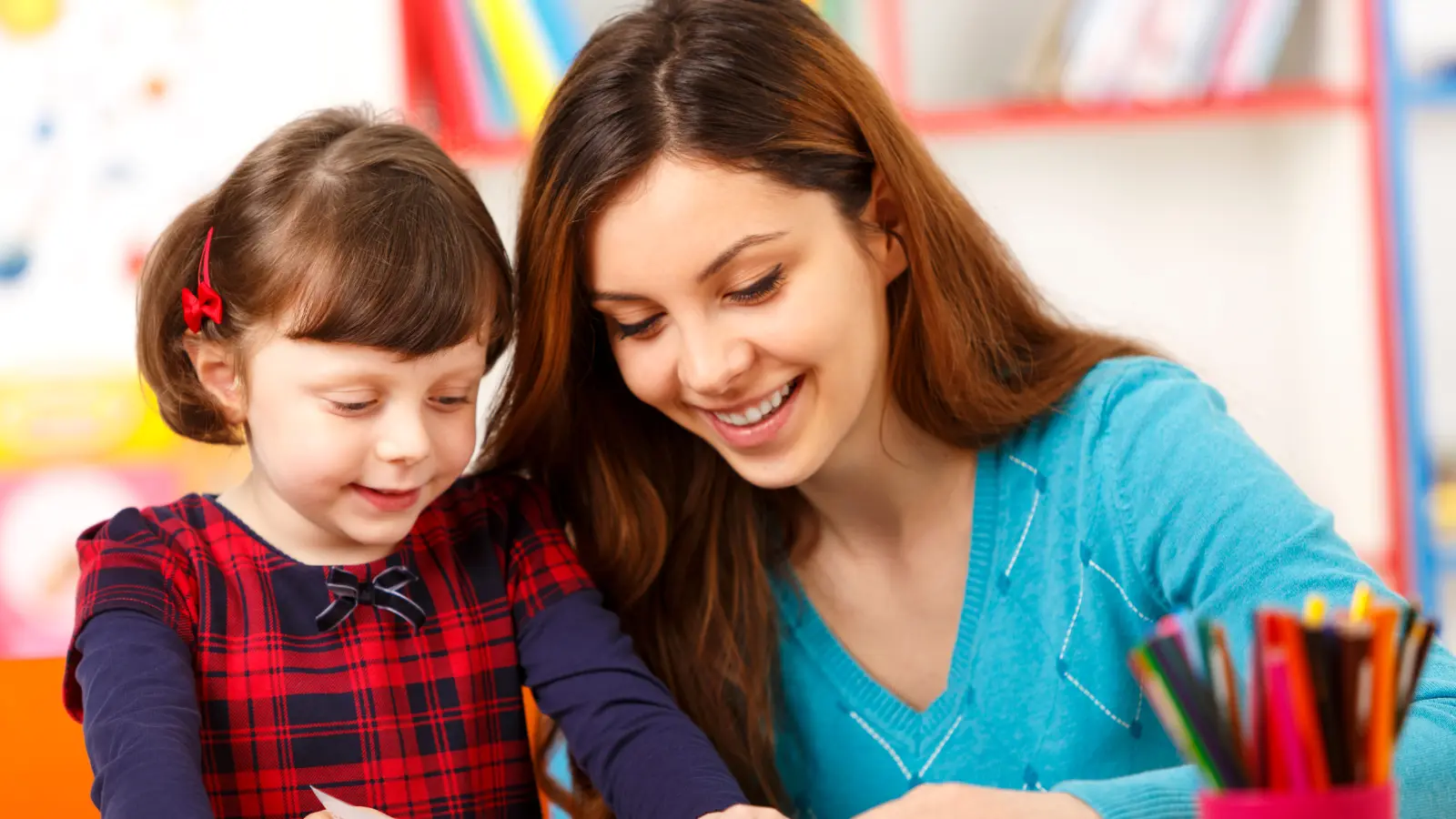 Adult and young girl smile while drawing at a table with coloured pencils.