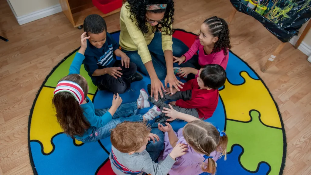 Adult and six children sit on a colouful rug with hands reaching to the centre.