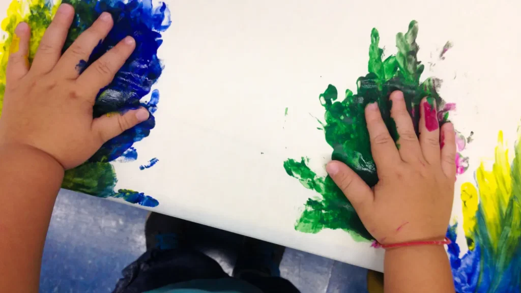 Child’s hands make green and blue handprint paint marks on white paper.