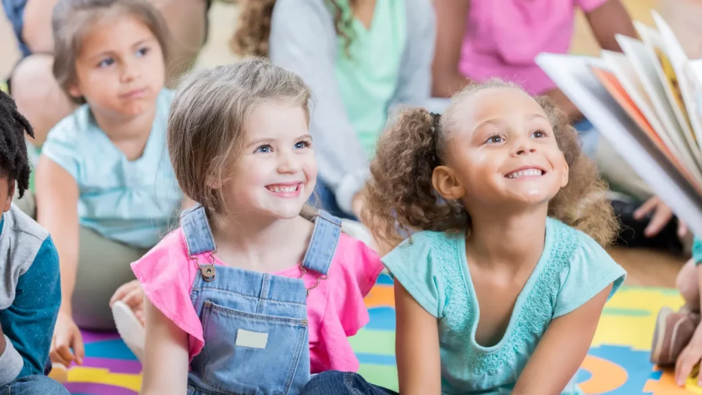 Two girls smile during story time while an adult reads to children.