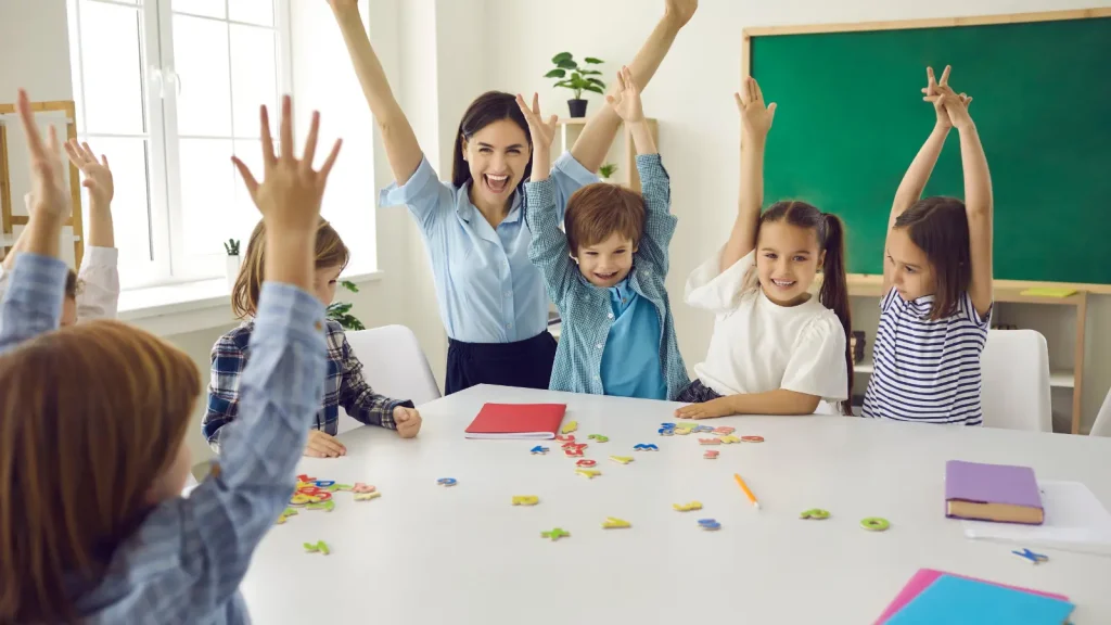 Adult and children raise their hands around a classroom table with letter tiles.