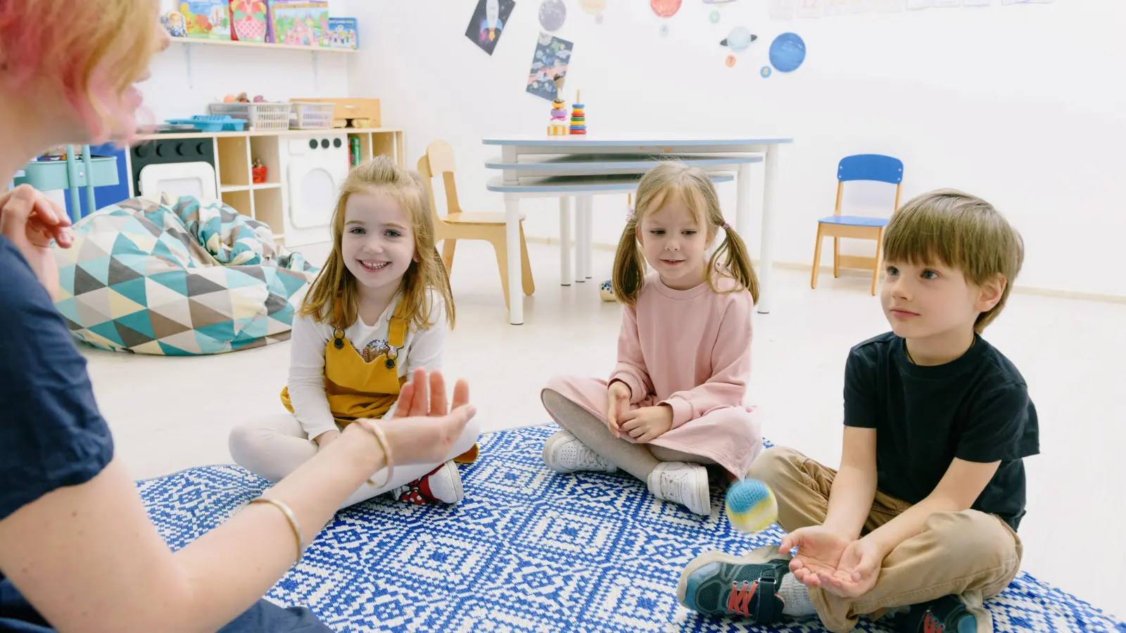 Adult leads a ball game with three children seated on a mat in a bright classroom.