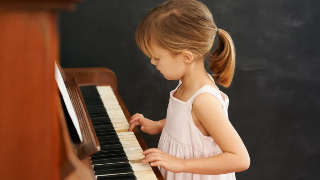 Young girl plays piano keys while standing beside an upright piano.