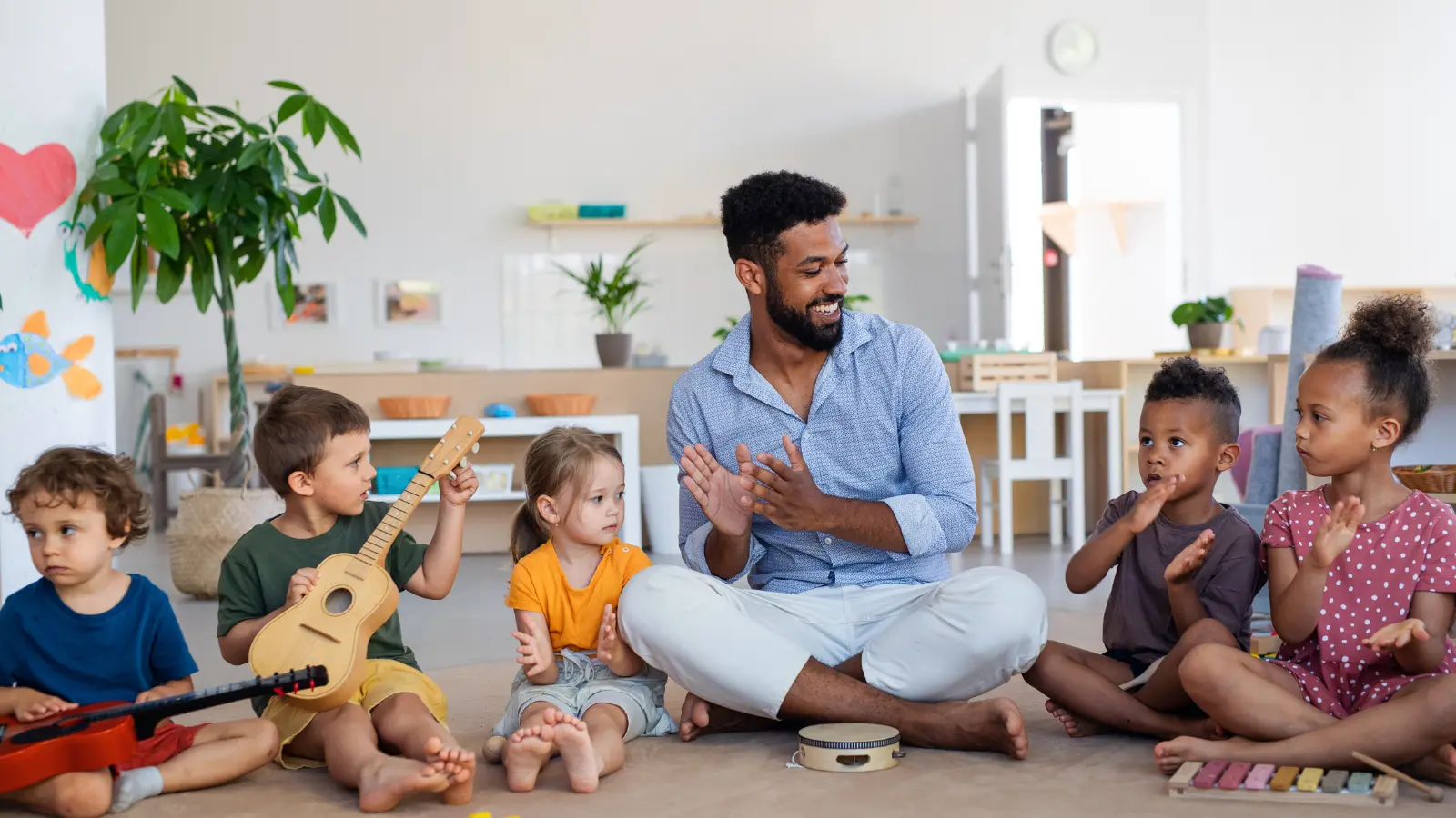 Adult sits with five children in a classroom, clapping while one child holds a ukulele.