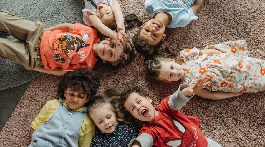 Seven children lie on a carpet in a circle, smiling up at the camera.