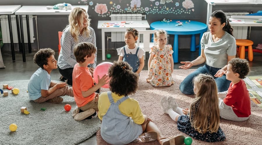 Two adults lead children in a circle activity on a nursery classroom carpet.