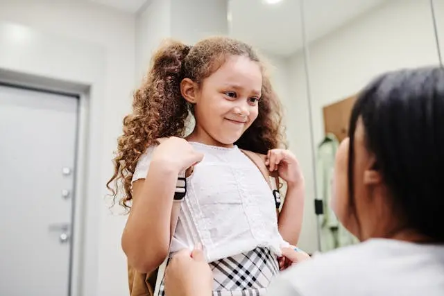 Child smiling while adult helps adjust backpack straps, getting ready to leave home