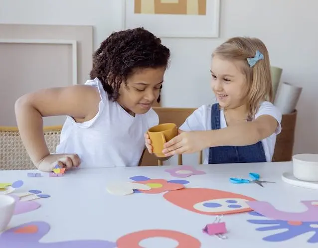 Two children doing arts and crafts at a table, smiling and sharing supplies