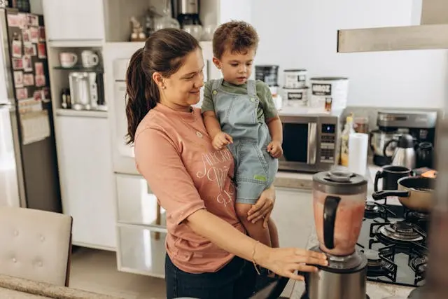 Adult holds a toddler in a kitchen while blending a drink on the counter.